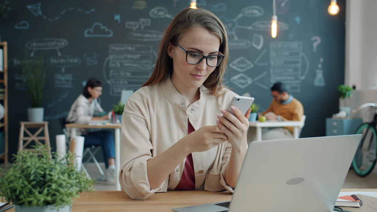 Seorang perempuan membalas pesan di ponsel sambil laptopnya terbuka di meja