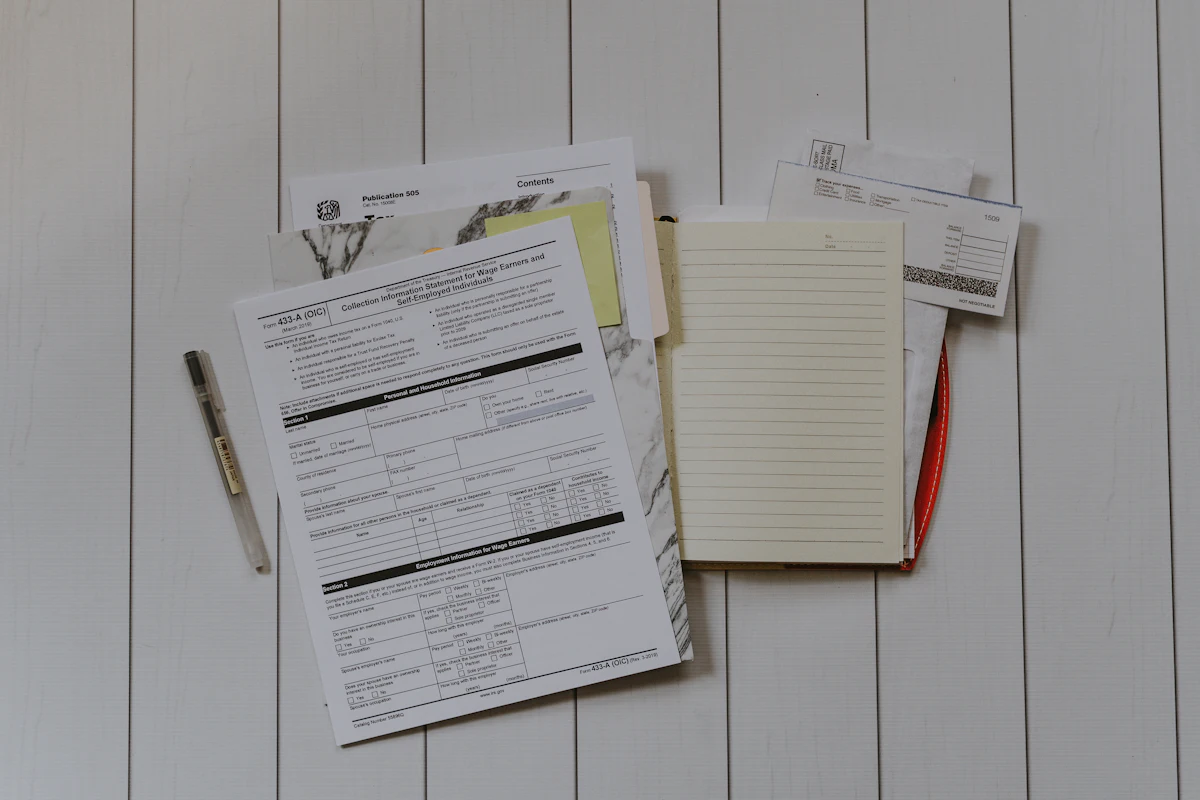 Dokumen dan formulir bertumpuk di meja kerja dengan sticky note kuning, buku catatan terbuka, dan pulpen