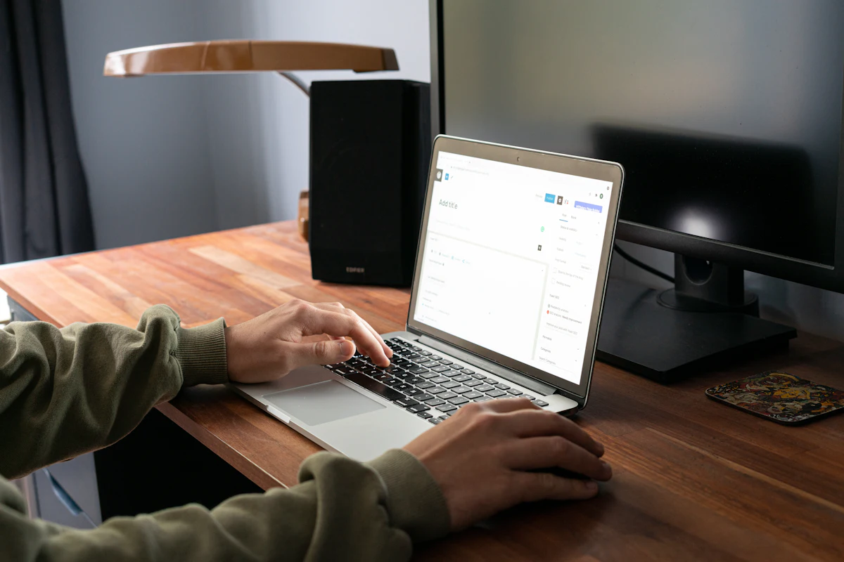 Tangan mengetik di laptop di meja kayu dengan cahaya jendela