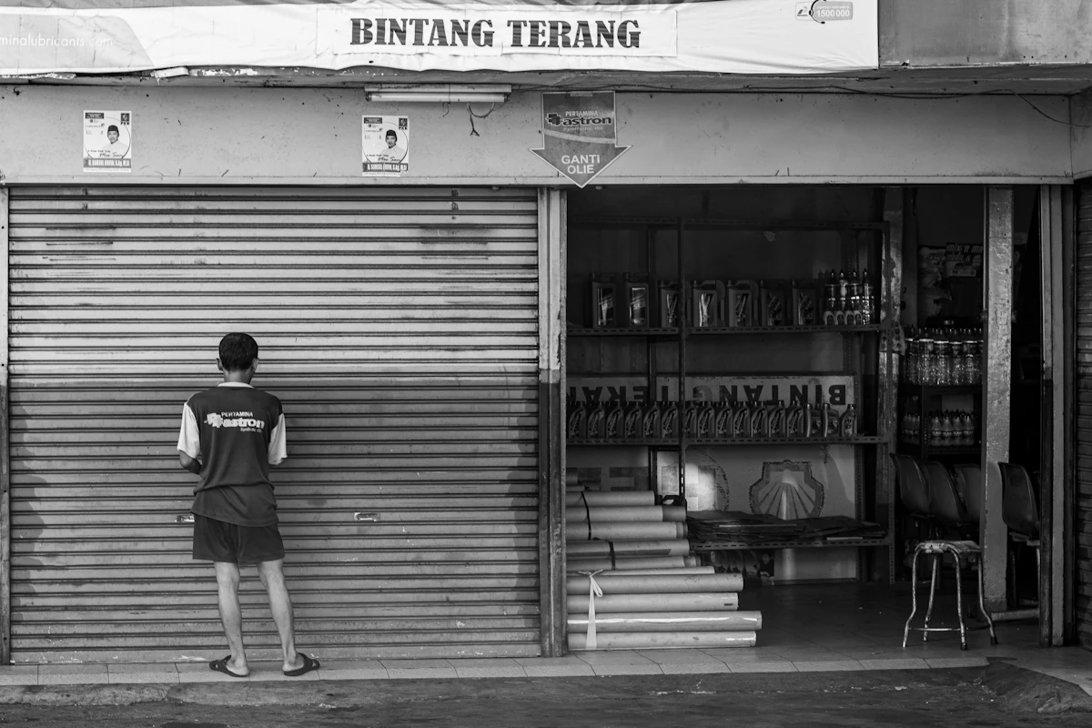 Seorang laki-laki berdiri di depan rolling door toko yang sudah tertutup, foto hitam putih