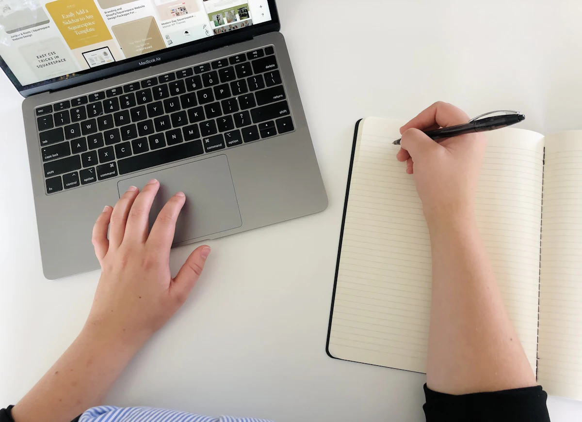 Seseorang menulis di buku catatan sambil membuka laptop di sampingnya