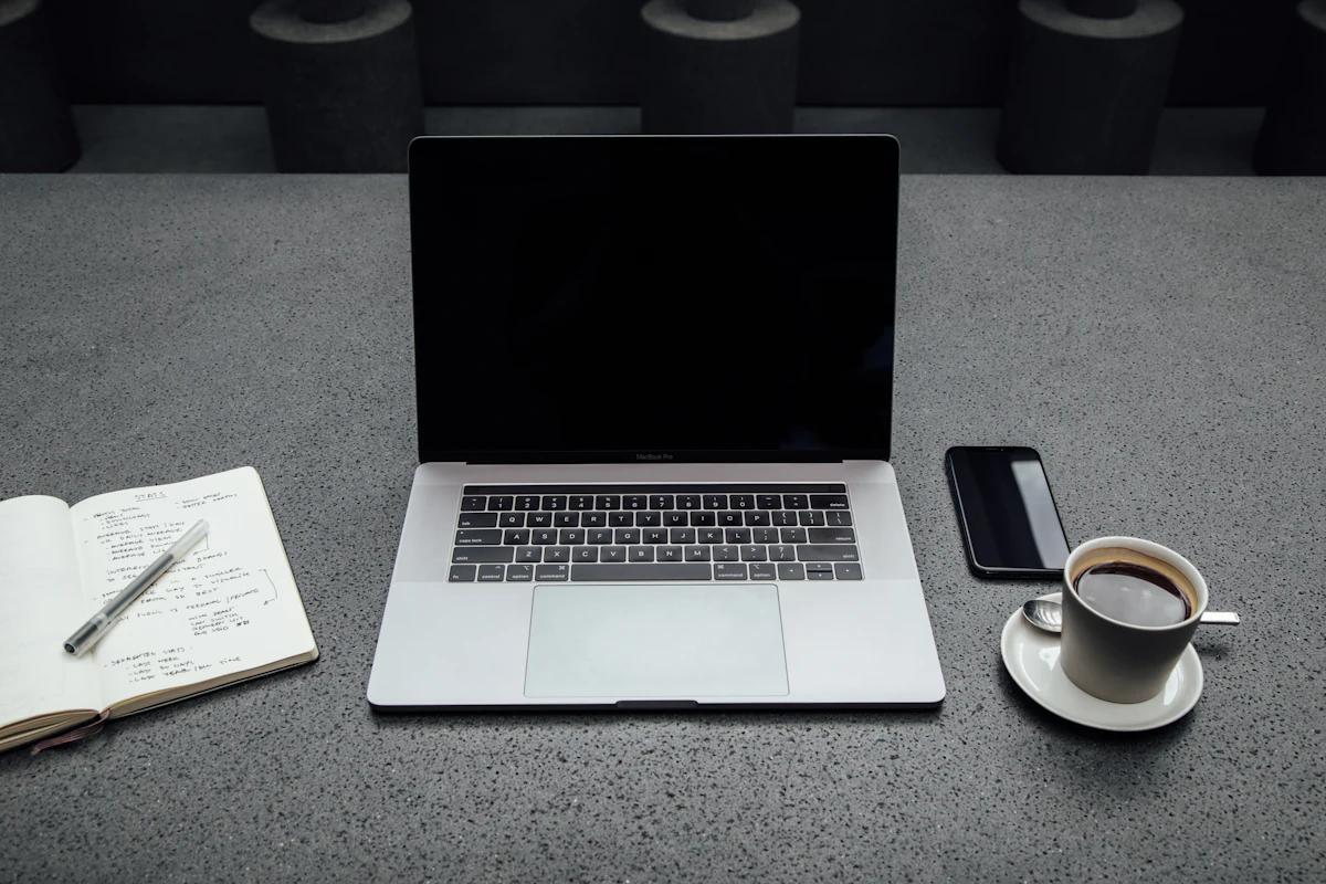 Meja kerja dengan secangkir kopi, ponsel, buku catatan terbuka berisi tulisan tangan, dan laptop yang tidak menyala