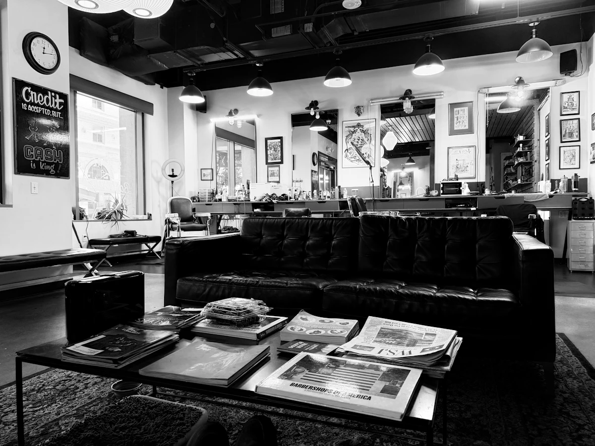 Sudut ruang tunggu sebuah barbershop dengan sofa dan tumpukan majalah, tidak ada pelanggan yang sedang menunggu