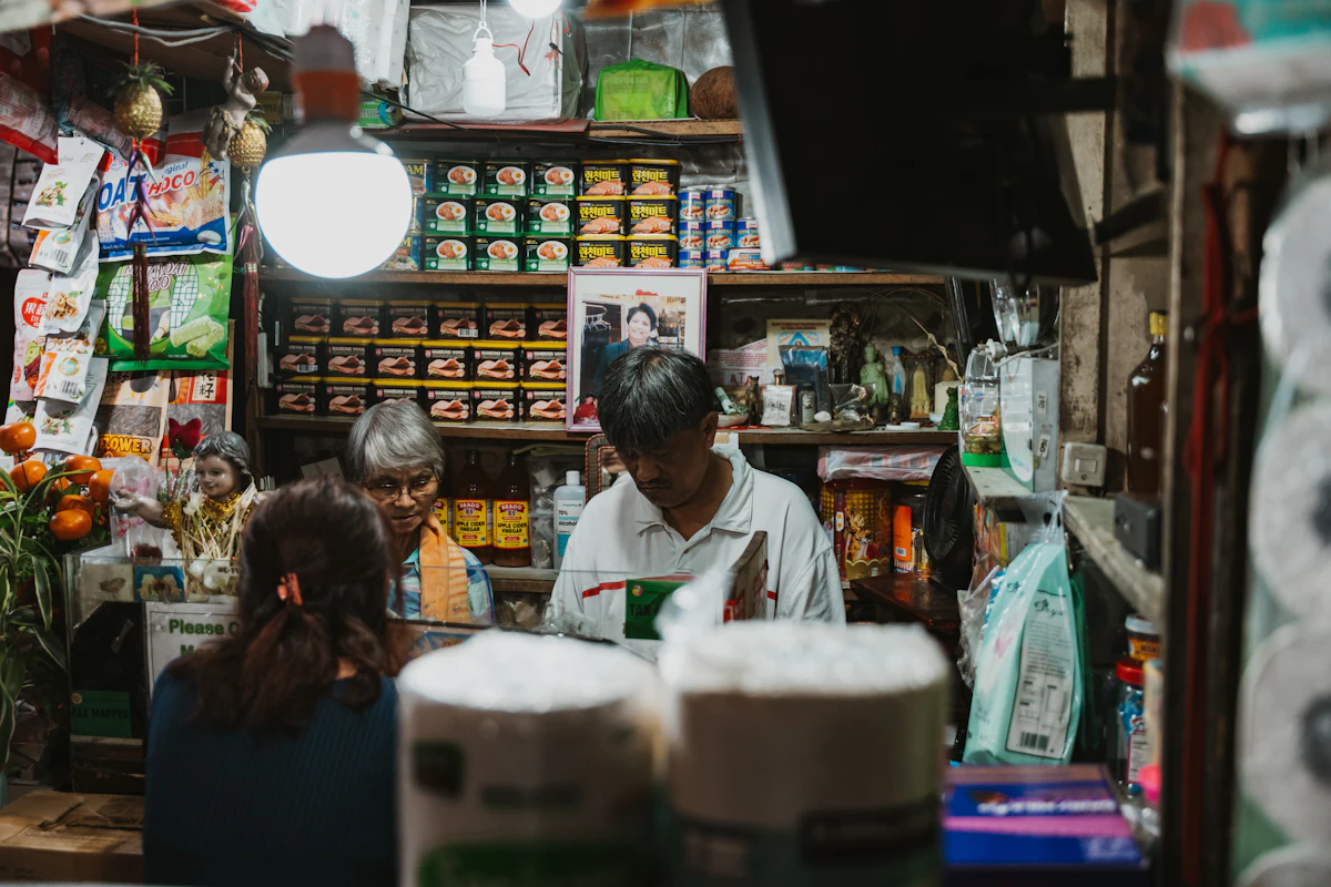 Dua pengelola toko bekerja berdampingan di balik konter yang penuh barang, dengan kardus dan tumpukan paket di sekitarnya