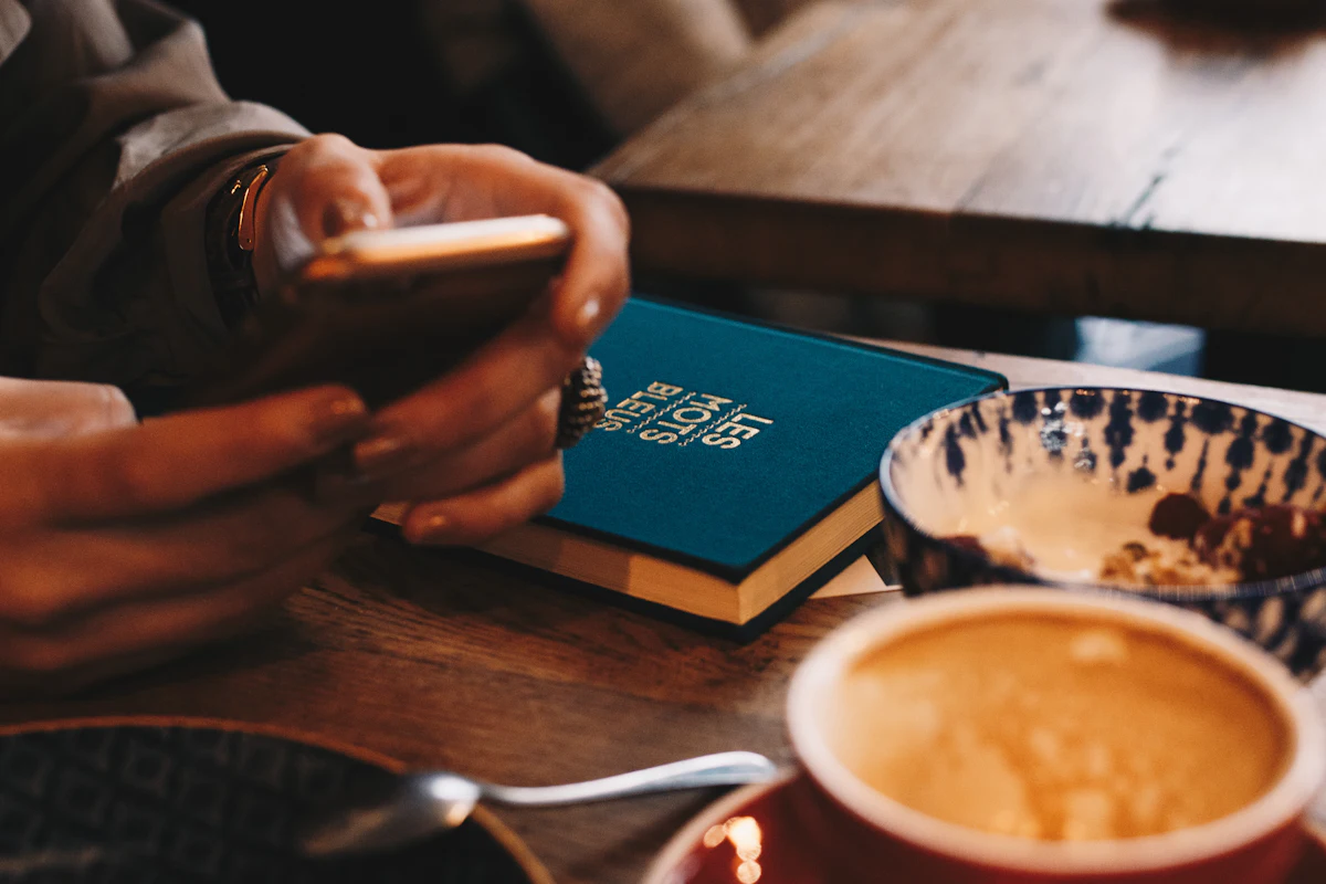 Dua tangan memegang ponsel di atas meja kayu, di sampingnya buku catatan dan cangkir kopi