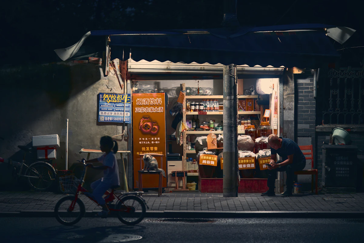 Sebuah toko kelontong kecil masih menyala pada malam hari, dengan seorang anak melintas bersepeda di jalan yang sudah sepi.