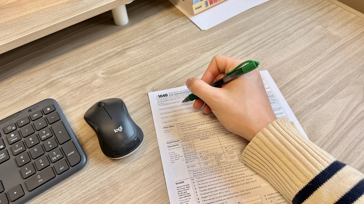 Tangan mengisi dokumen dengan pulpen di meja kerja, di samping keyboard dan mouse