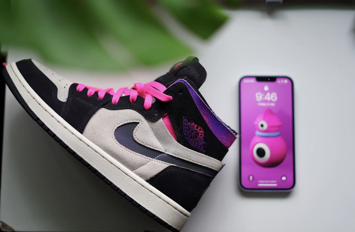 Sepatu sneaker dipajang di sebelah ponsel yang berdiri di atas latar netral untuk pemotretan produk katalog