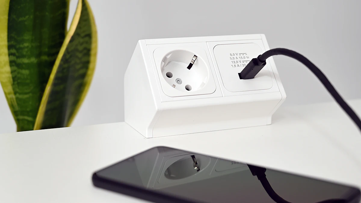 Ponsel tergeletak mengisi daya lewat kabel USB-C dari stopkontak meja di atas meja putih