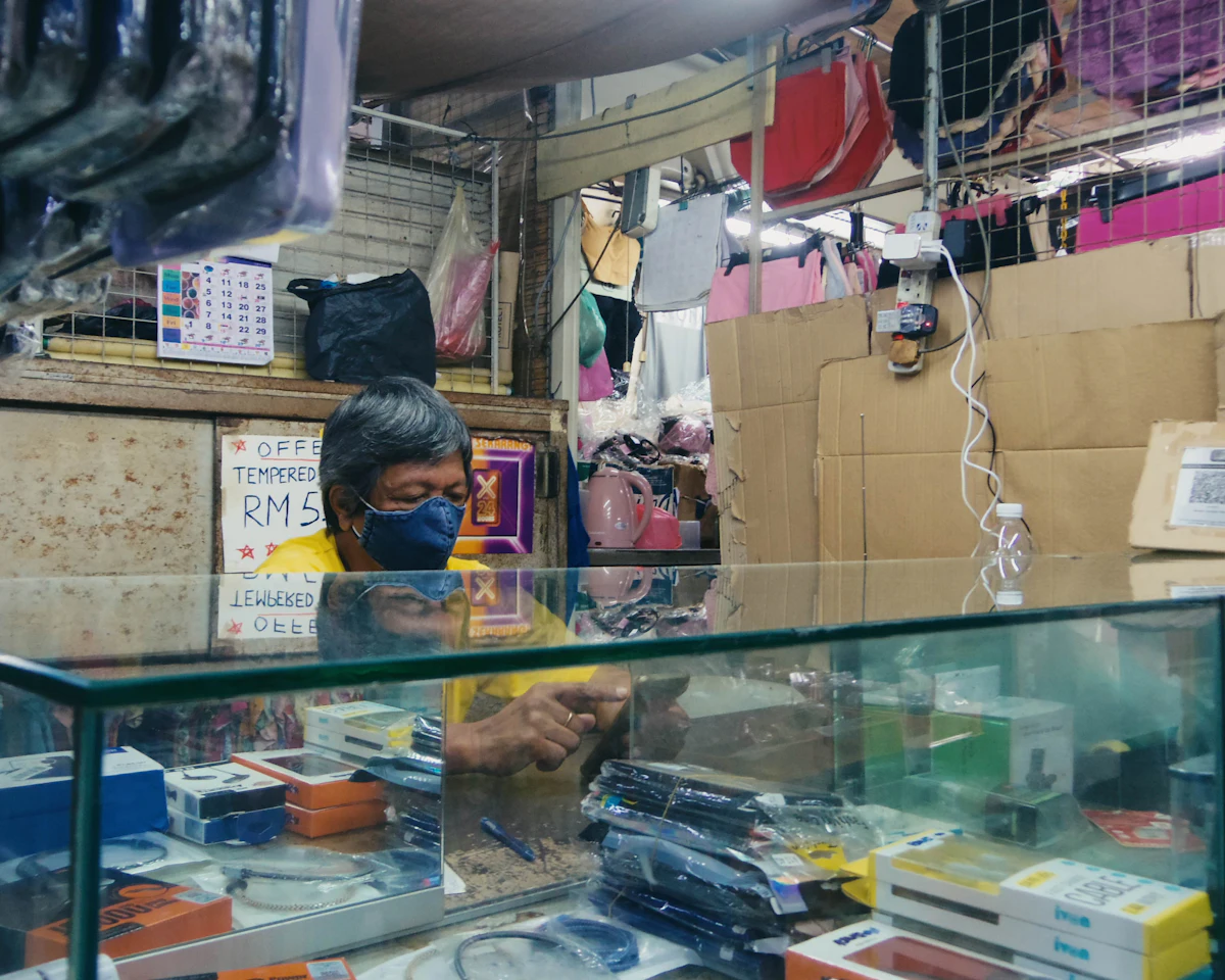 Penjual toko kecil bermasker memegang ponsel di balik meja kasir kaca pada siang hari, kardus tertumpuk di sekitar kiosnya