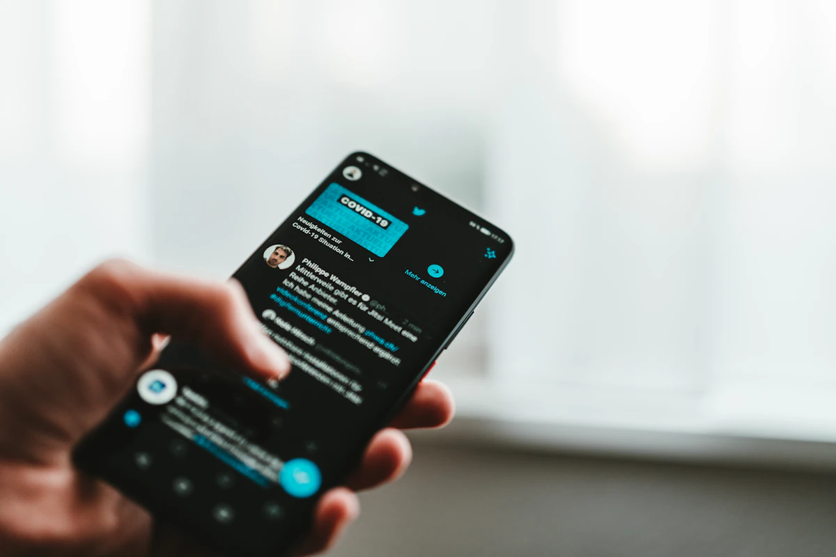Tangan memegang ponsel dengan utas percakapan dan papan ketik terbuka di layar, jempol di dekat keyboard untuk mengetik balasan, latar hijau luar ruangan tampak kabur