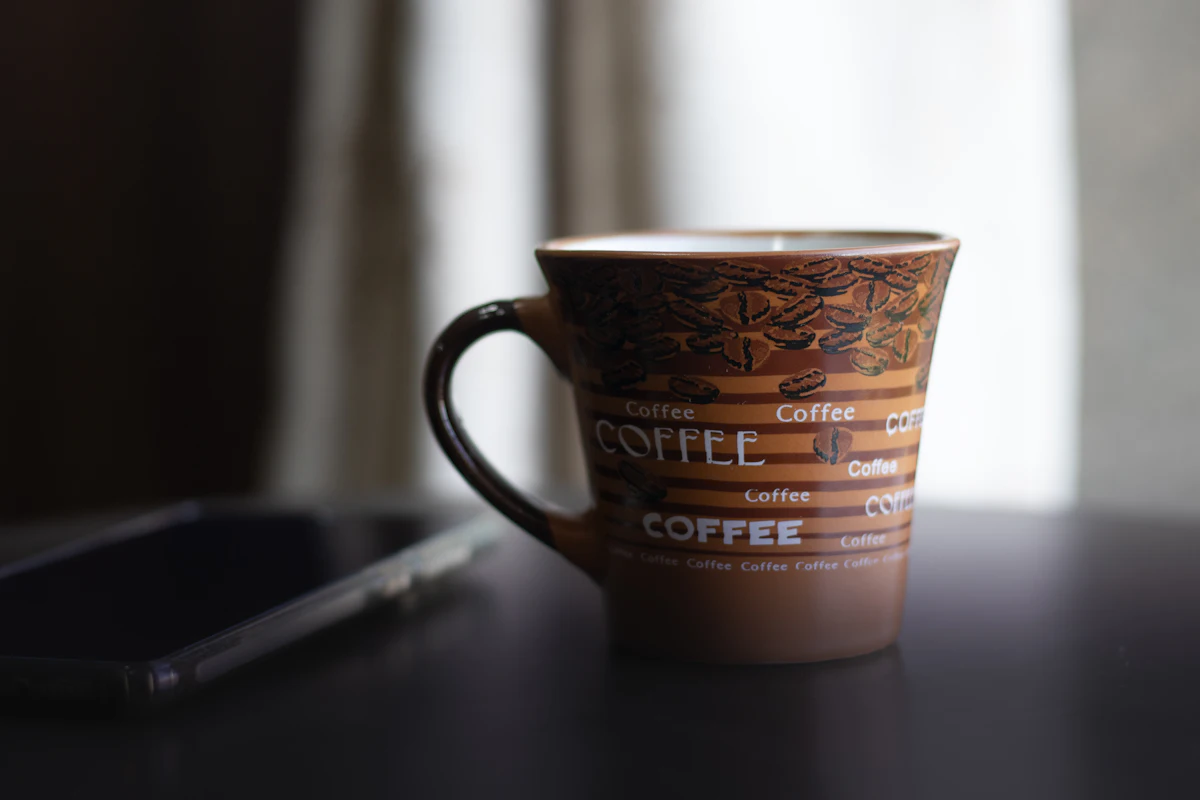 secangkir kopi dan ponsel tergeletak di meja dengan cahaya pagi dari jendela
