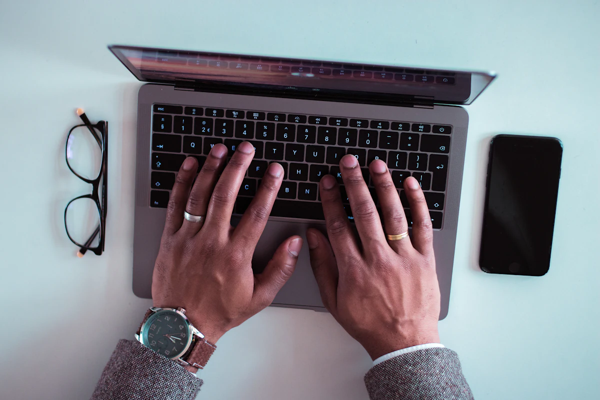 Tangan mengetik di keyboard laptop, ponsel tergeletak di sebelah kanan dan kacamata di sebelah kiri