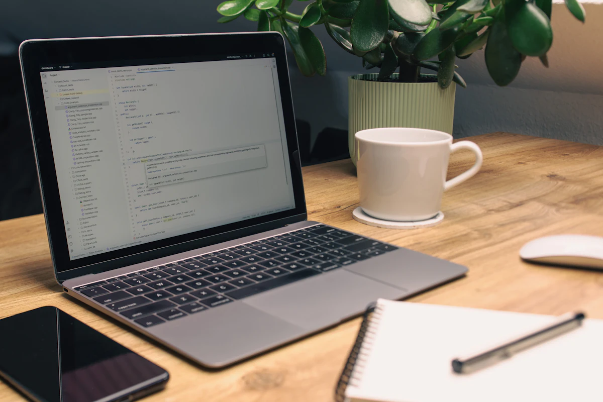Laptop terbuka di meja bersama buku catatan, ponsel, dan cangkir kopi