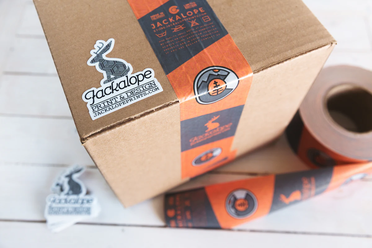 Kardus paket cokelat dengan stiker logo hitam di bagian penutupnya, siap dikirim