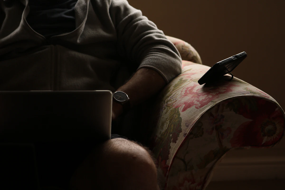Seseorang duduk di kursi rumah memegang ponsel dengan laptop tergeletak di sampingnya di luar jam kerja