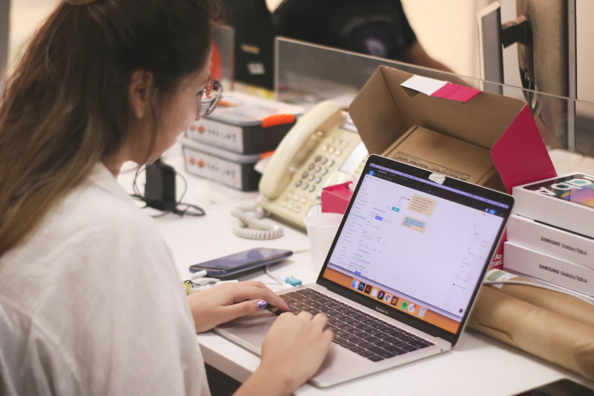 Pemilik toko mengetik di laptop di meja kerja yang dipenuhi kardus paket terbuka dan tumpukan kotak, dengan ponsel tergeletak di sampingnya