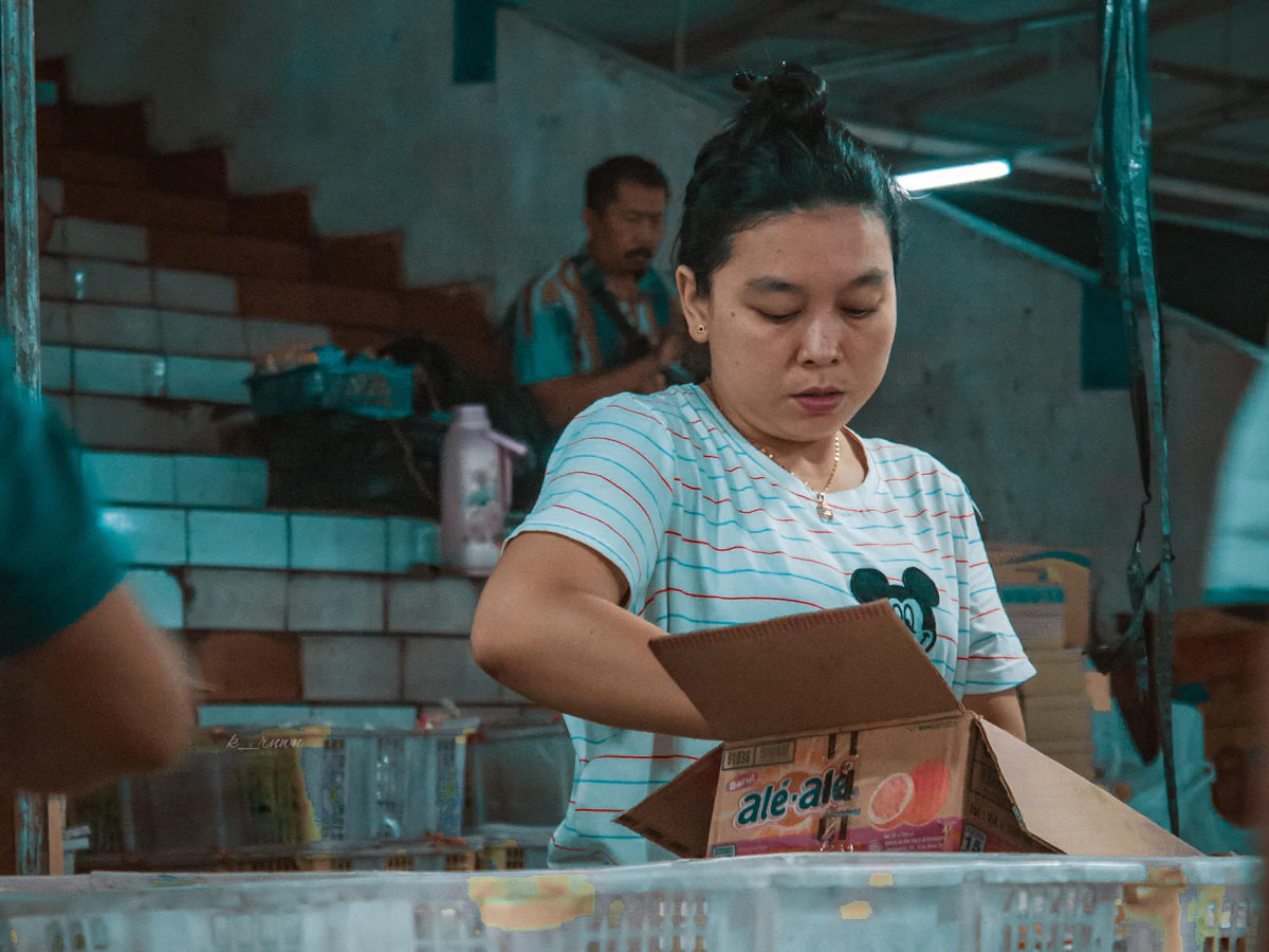 Seorang perempuan berkaus garis merah putih memegang kardus paket di meja packing